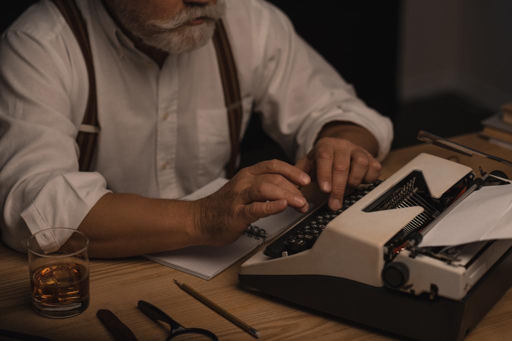 обрезанный снимок старшего писателя, работающего с пишущей машинкой, изолированный на черном
 - Фото, изображение