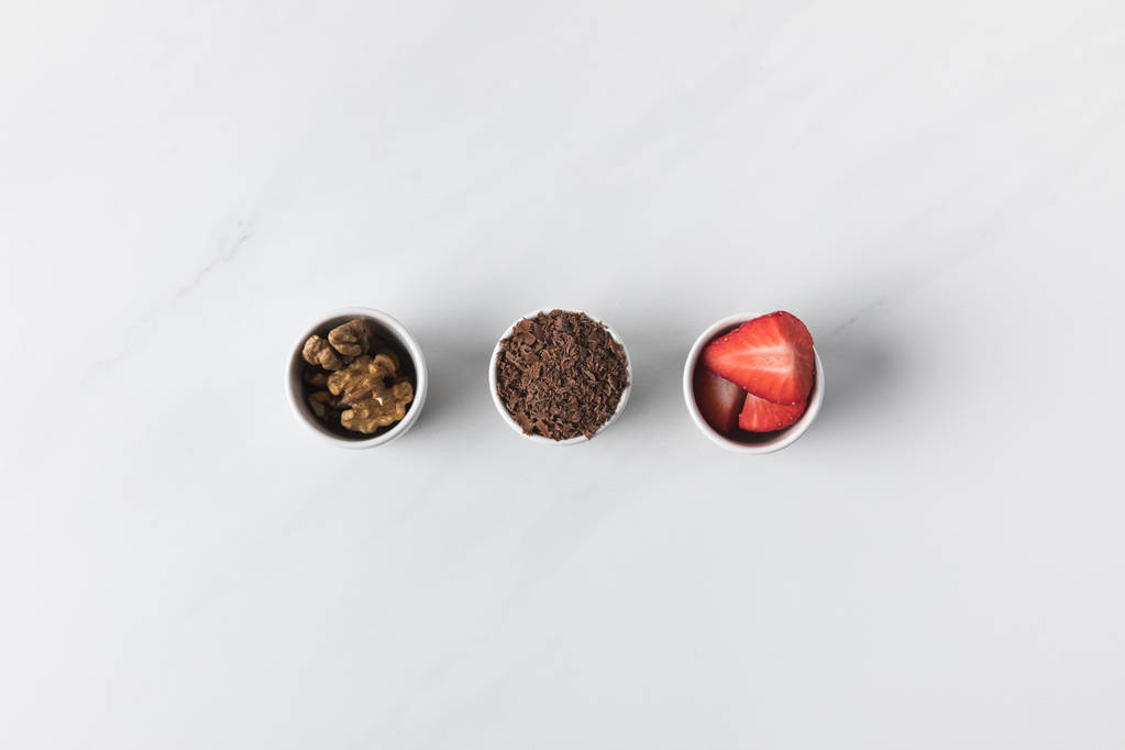 Kolme kulhoa saksanpähkinöitä, raastettua suklaata ja mansikoita valkoisella
 - Valokuva, kuva