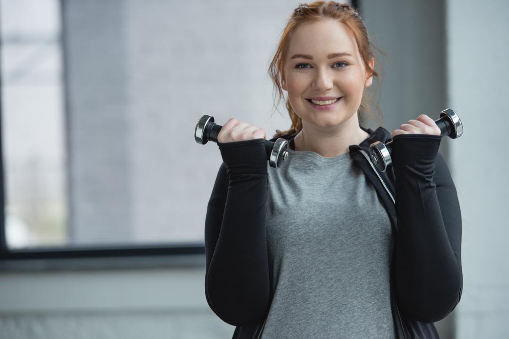 Chica obesa sonriente entrenando con mancuernas en el gimnasio
 - Foto, Imagen