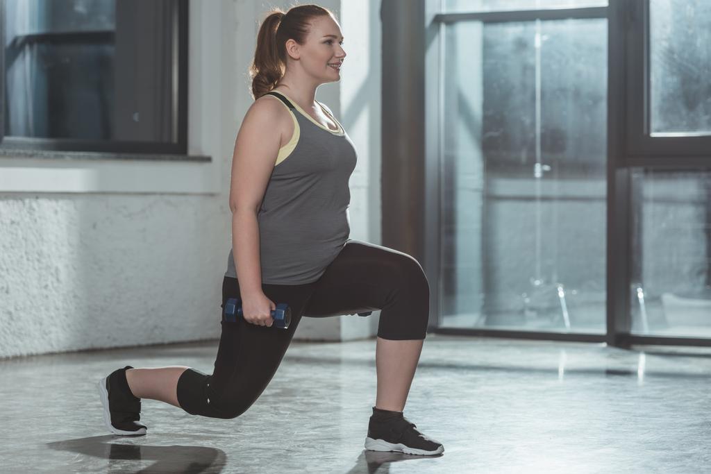 Chica con sobrepeso realizando embestidas con mancuernas en el gimnasio
 - Foto, Imagen