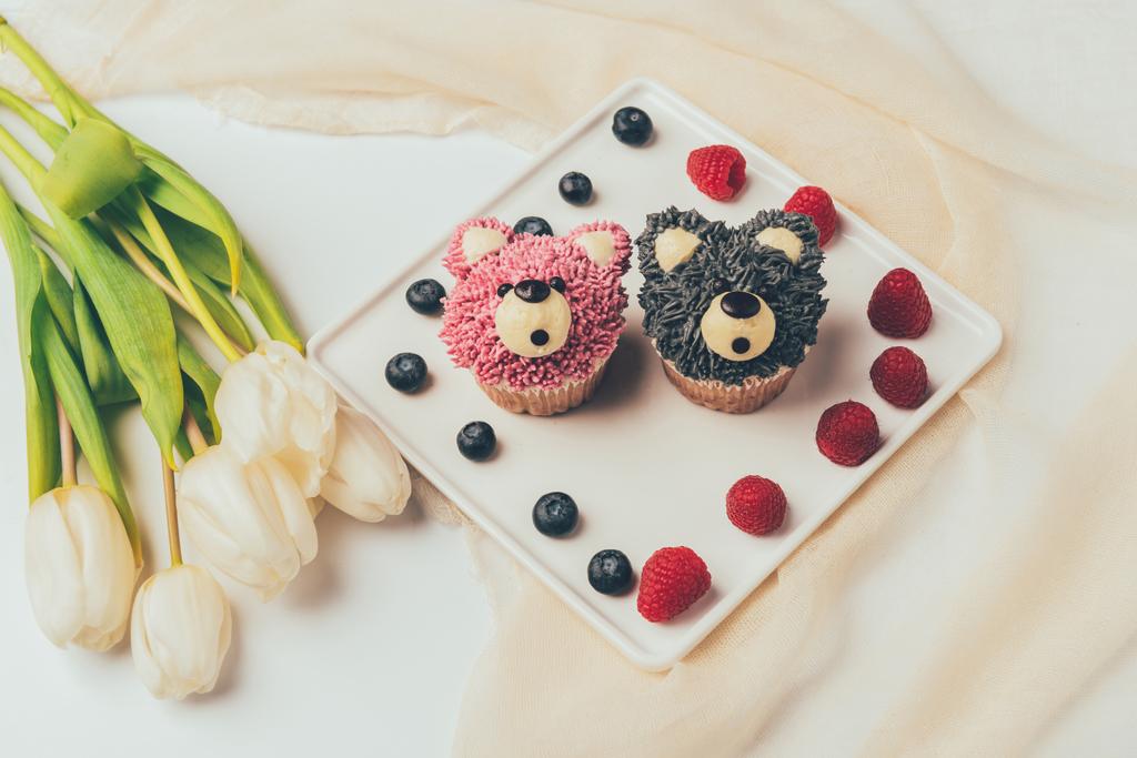 Nahaufnahme köstlicher Muffins in Form von Bären, frischen Beeren und Tulpenblüten  - Foto, Bild