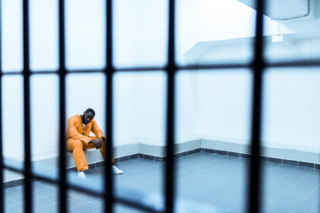 Αφρικανική αμερικανική αιχμάλωτος που κάθεται στον πάγκο πίσω από τα κάγκελα της φυλακής - Φωτογραφία, εικόνα