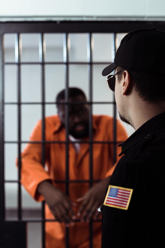 Σεκιουριτάς, στέκεται κοντά φυλακής τα σίδερα και κοιτάζοντας αφρικανική αμερικανική αιχμάλωτος  - Φωτογραφία, εικόνα