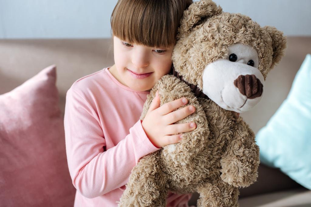 Kind mit Down-Syndrom sitzt auf Sofa und umarmt Teddybär - Foto, Bild