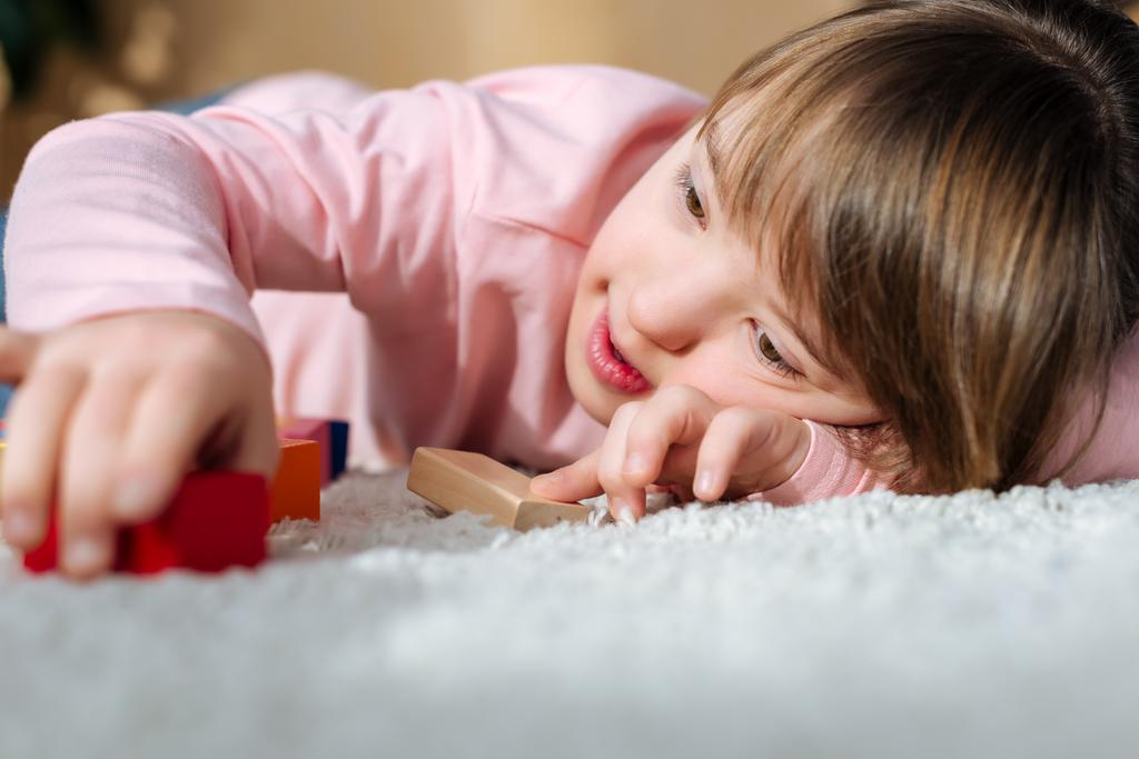 Ребенок с синдромом Дауна играет с кубиками игрушек, лежа на полу
 - Фото, изображение