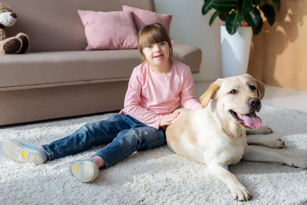 Niño con síndrome de Down sentado en el suelo con Labrador retriever en la alfombra
  - Foto, Imagen