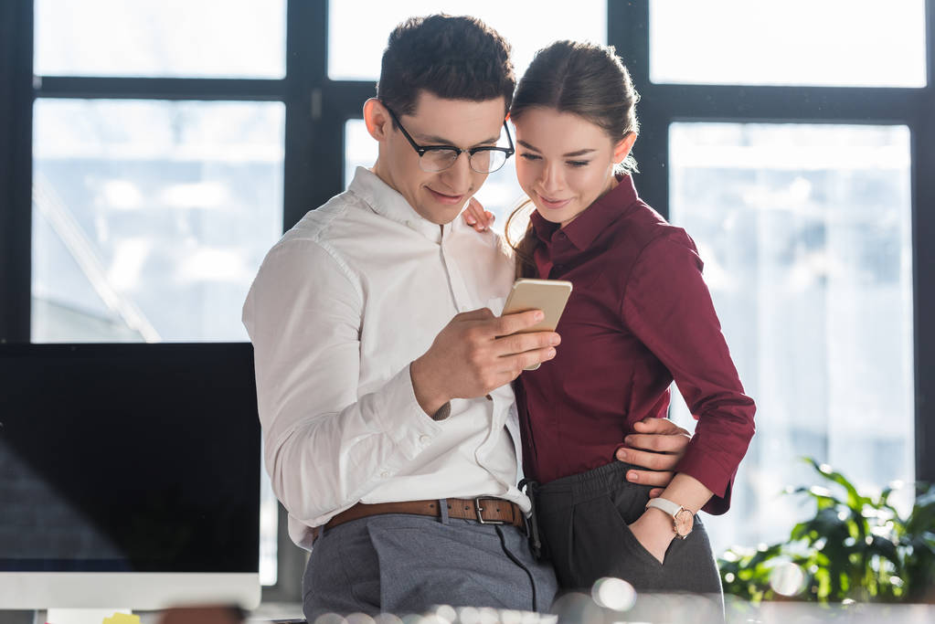 attractive young businesspeople in formal clothing having office romance and using smartphone together - Photo, Image