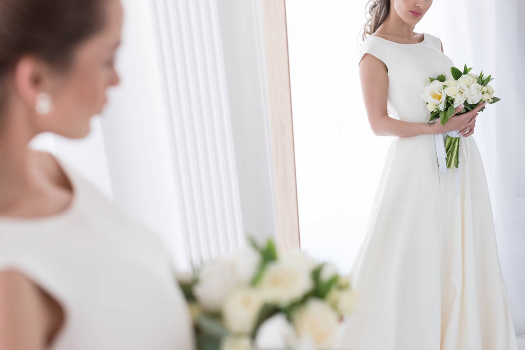 bride in traditional dress with wedding bouquet looking at her reflection in mirror - Photo, Image