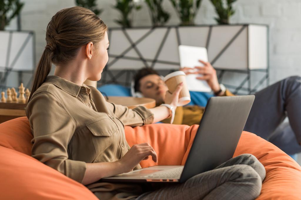 mujer joven beber café y trabajar con el ordenador portátil mientras está sentado en la bolsa de frijoles y taza de paso a novio
 - Foto, Imagen