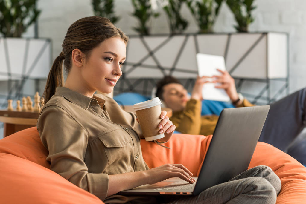 young woman drinking coffee and working with laptop while sitting in bean bag - Photo, Image