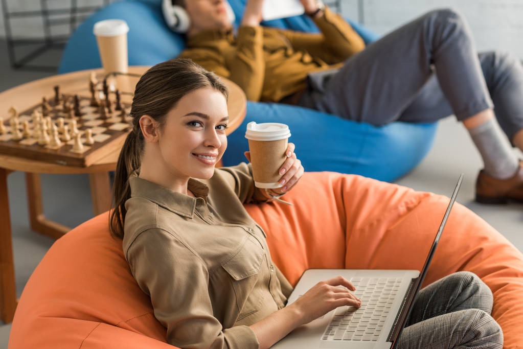щаслива молода жінка п'є каву і працює з ноутбуком, сидячи в бобовій сумці
 - Фото, зображення