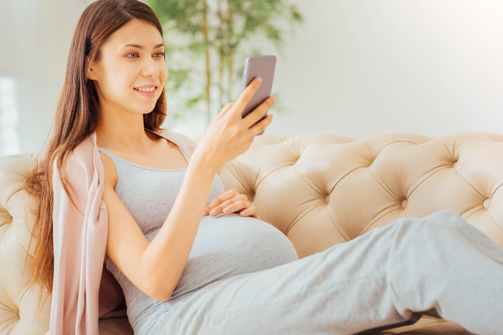 Dość rodząca kobieta przy użyciu smartfona, leżąc na kanapie. - Zdjęcie, obraz