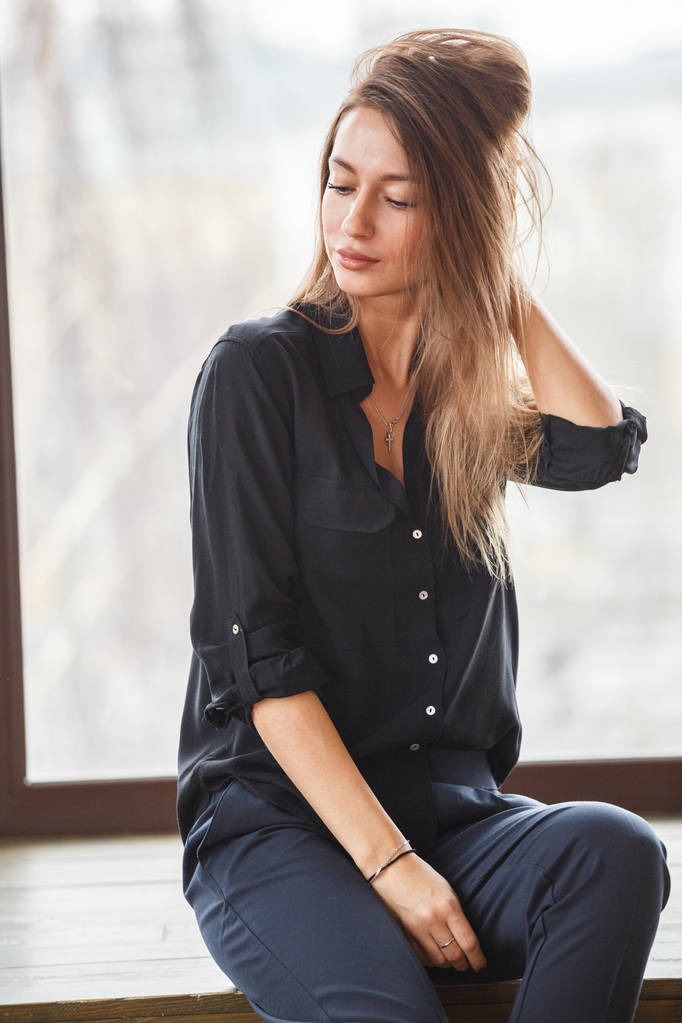 Studio shot di bella giovane donna vestita con abiti casual neri seduta sul pavimento in legno, finestre illuminate sfondo
 - Foto, immagini