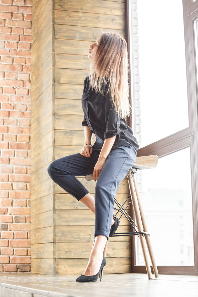 Studio βολή του όμορφη νεαρή γυναίκα που φοράει μαύρο casual ρούχα που κάθεται σε ξύλινη καρέκλα, φόντο φωτισμένο παράθυρο - Φωτογραφία, εικόνα