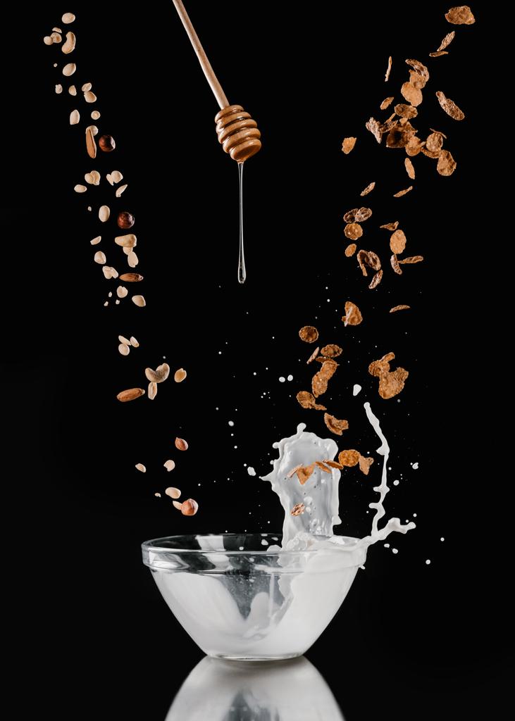 caída de nueces y copos de maíz y goteo de miel en un tazón con leche en negro
 - Foto, Imagen
