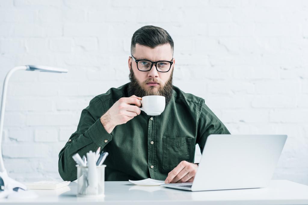 portrait of serious businessman with cup of coffee at workplace with laptop - Photo, Image
