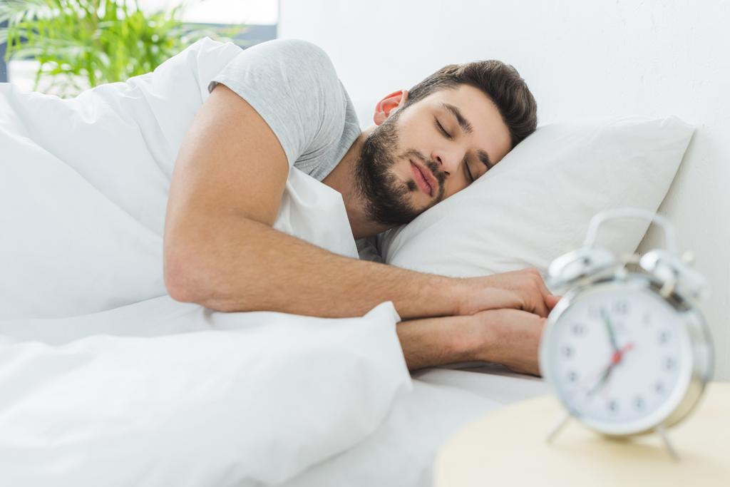 hezký vousatý muž spí v posteli ráno, budík na popředí - Fotografie, Obrázek