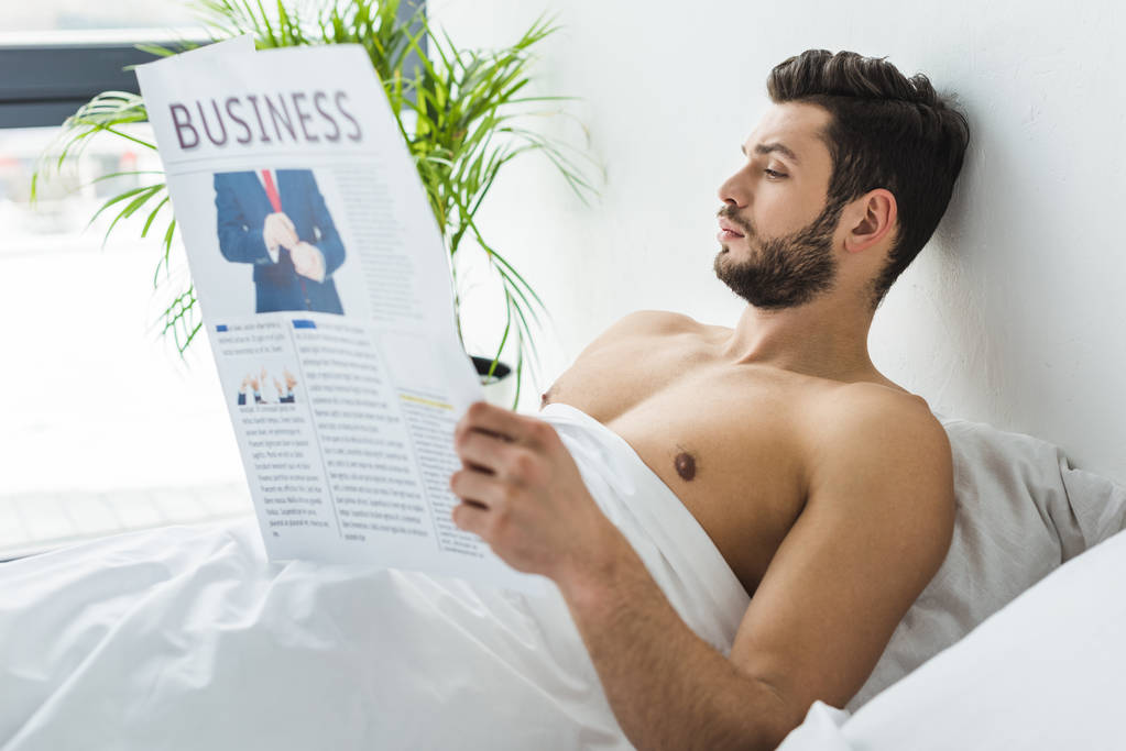 上半身裸のひげを生やした男が朝ベッドで新聞を読む - 写真・画像