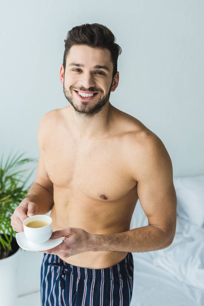 hombre feliz sin camisa de pie en el dormitorio con una taza de café
 - Foto, Imagen