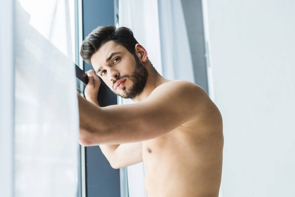 Hemdloser Mann steht am Fenster und blickt in Kamera - Foto, Bild