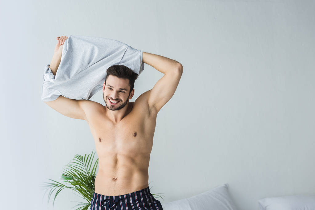 przystojny seksowny uśmiechający się człowiek startu jego t-shirt w sypialni - Zdjęcie, obraz