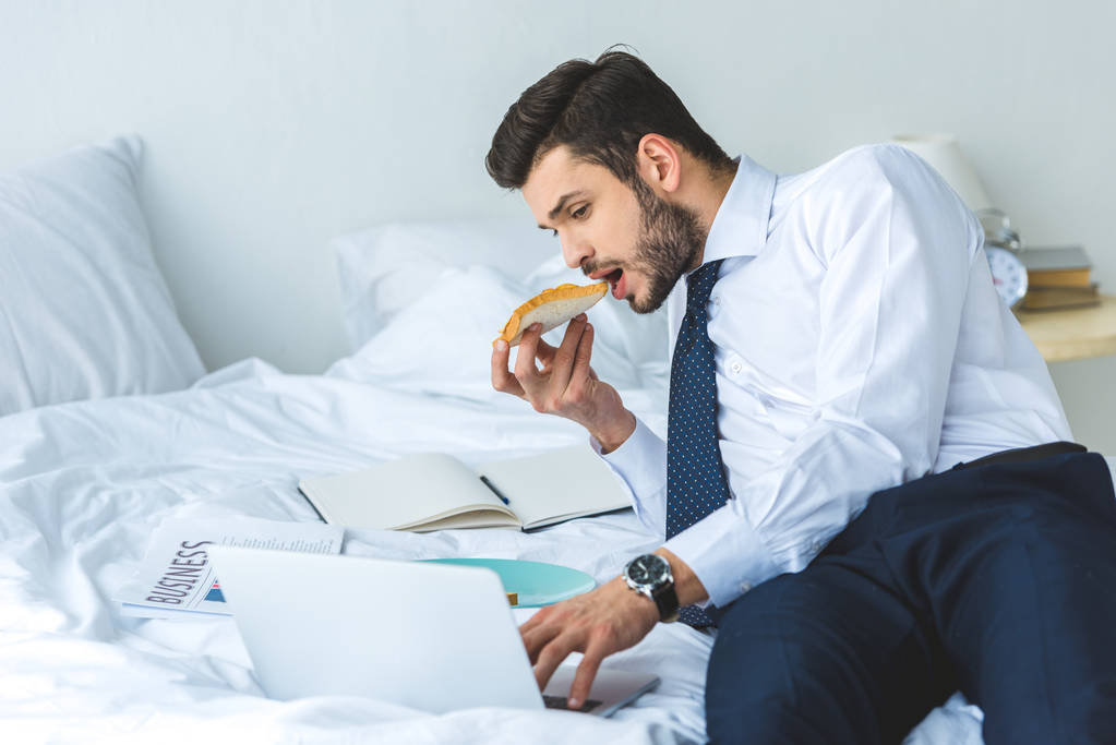Geschäftsmann isst Toast zum Frühstück, während er mit Laptop im Bett arbeitet - Foto, Bild