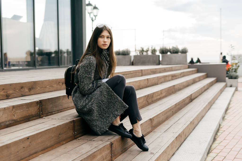stylische junge dunkelhaarige Mädchen in einem grauen Mantel sitzt auf den Stufen in den Straßen der Stadt und posiert - Foto, Bild