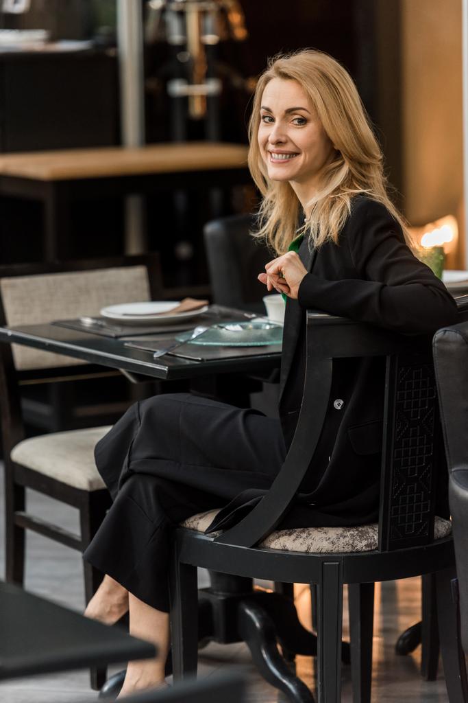 kaunis hymyilevä nainen katselee kameraa istuessaan pöydässä kahvilassa
 - Valokuva, kuva