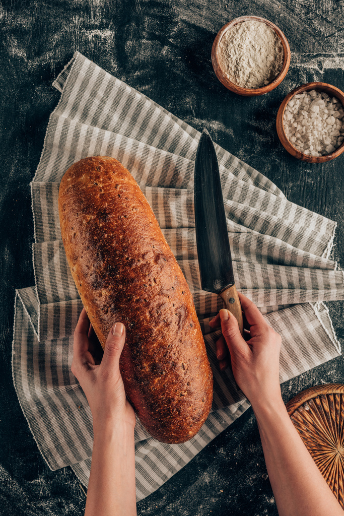 částečný pohled ženských rukou, nůž a bochník chleba na plátno na tmavý povrch s moukou - Fotografie, Obrázek