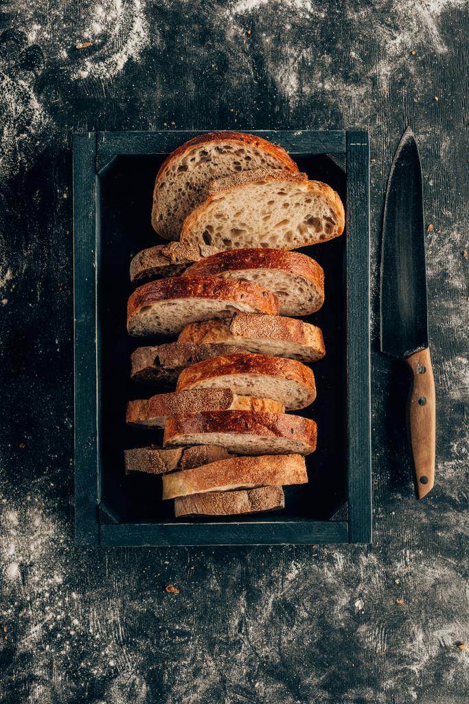 vista superior de piezas dispuestas de pan en caja de madera y cuchillo sobre mesa oscura con harina
 - Foto, Imagen