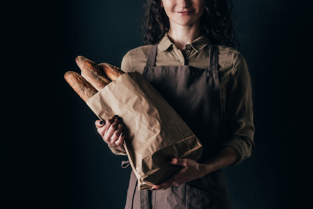 cropped shot of woman in apron holding french baguettes in paper bag in hands isolated on black - Photo, Image