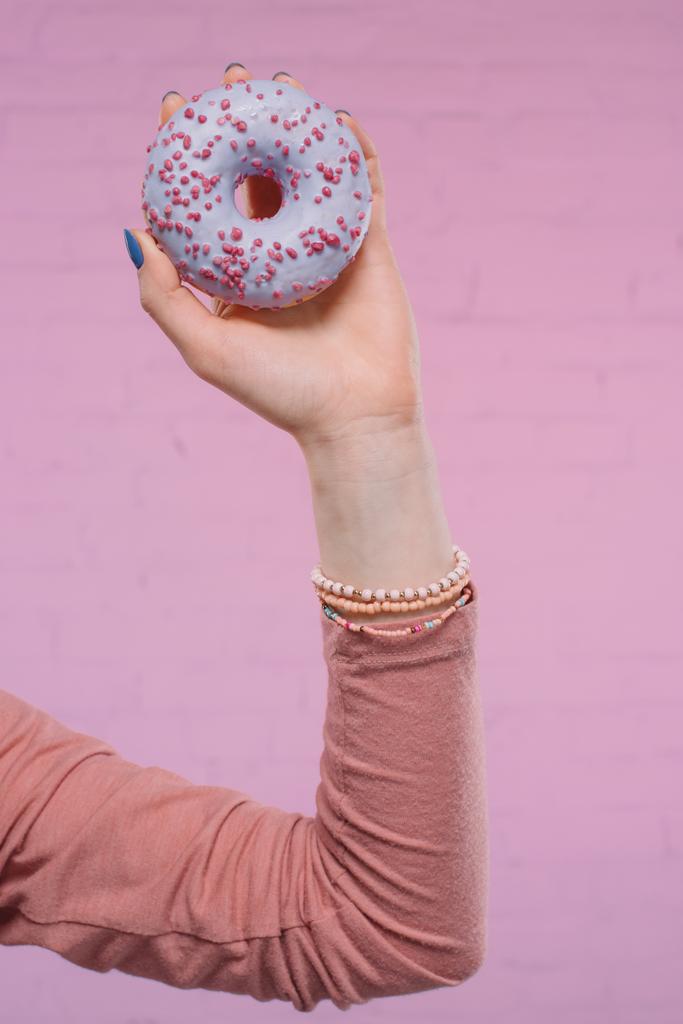 обрезанный снимок женщины с восхитительным глазурованным пончиком в руке перед розовой кирпичной стеной
 - Фото, изображение
