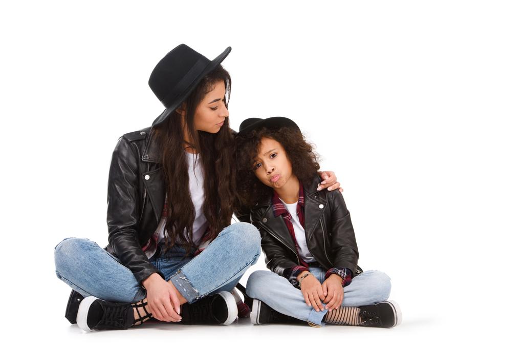 красива мати і дочка в шкіряних куртках і капелюхах сидять на підлозі ізольовані на білому
 - Фото, зображення