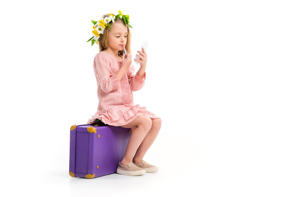 Widok z boku dziecko siedzi na fioletowy walizkę i robi makijaż przez szminka na białym tle - Zdjęcie, obraz