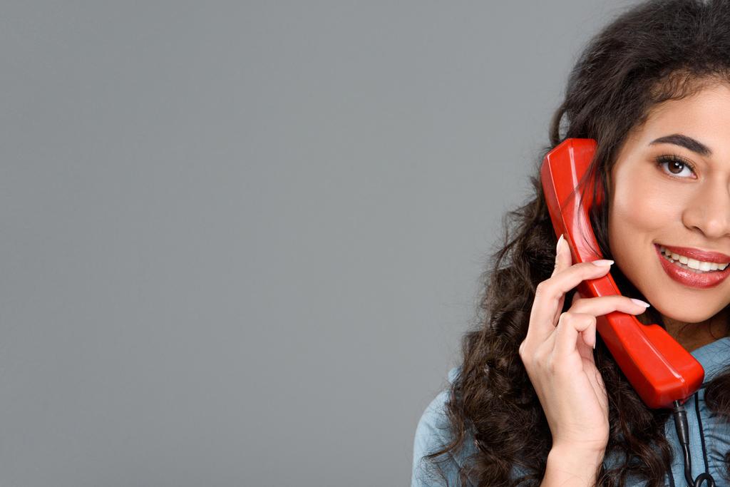 улыбающаяся молодая женщина, разговаривающая по старинному красному телефону, изолированному на сером
 - Фото, изображение