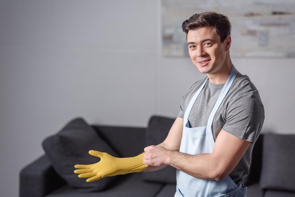 lachende knappe man dragen van rubberen handschoen voor het schoonmaken in woonkamer - Foto, afbeelding