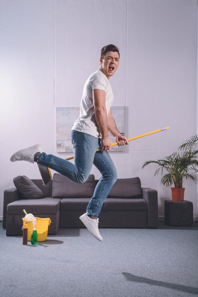 homem bonito pulando com vassoura na sala de estar
 - Foto, Imagem