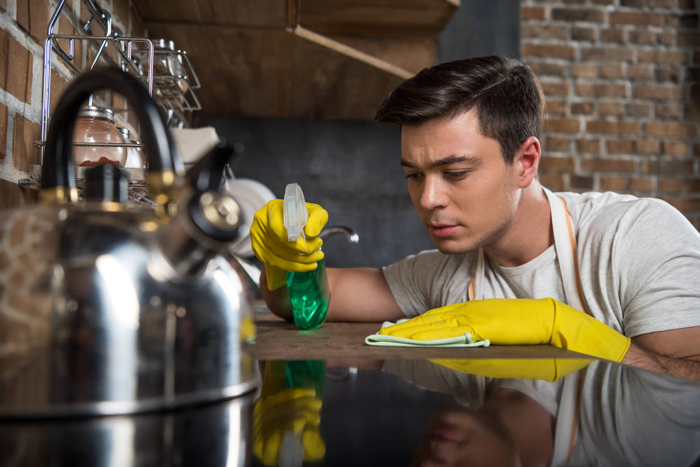 hombre guapo serio limpieza cocina con aerosol botella y trapo
 - Foto, Imagen