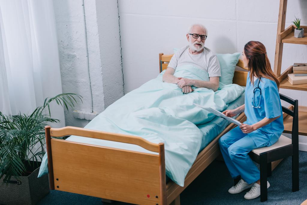 Vanha mies potilas ja sairaanhoitaja keskustelevat uutisia sanomalehdestä
 - Valokuva, kuva