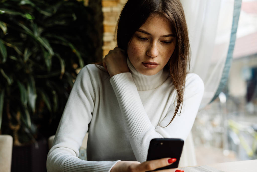 mladá černovlasá dívka v bílém svetru sedí v kavárně, na jídlo čeká a vypadá v ní smartphone - Fotografie, Obrázek