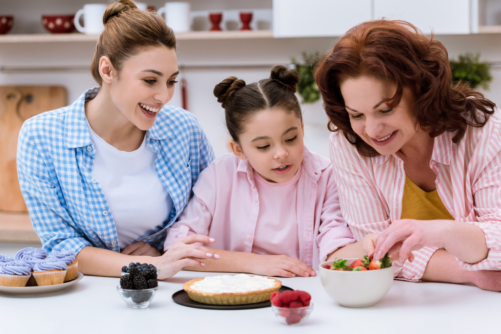 три поколения женщин вместе украшают десерт ягодами на кухне
 - Фото, изображение