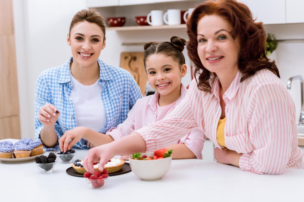 three generations of women decorating dessert with berries together at kitchen and looking at camera - Photo, Image