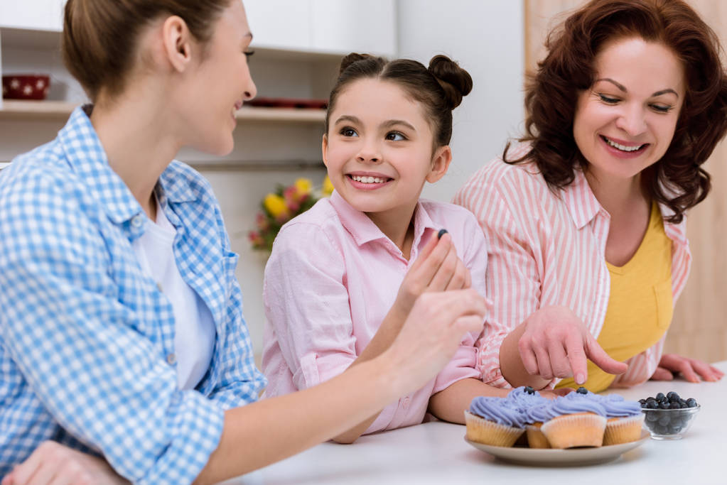 Drei Generationen von Frauen verzieren gemeinsam in der Küche Cupcakes mit Blaubeeren - Foto, Bild