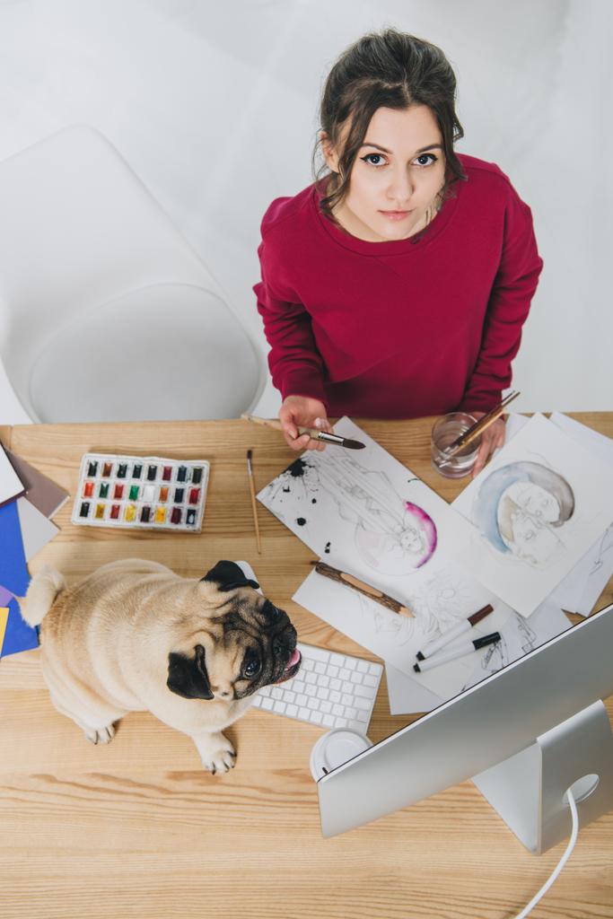 attraktive junge Mädchen arbeitet an Illustrationen mit niedlichen Mops auf Arbeitstisch mit Computer - Foto, Bild