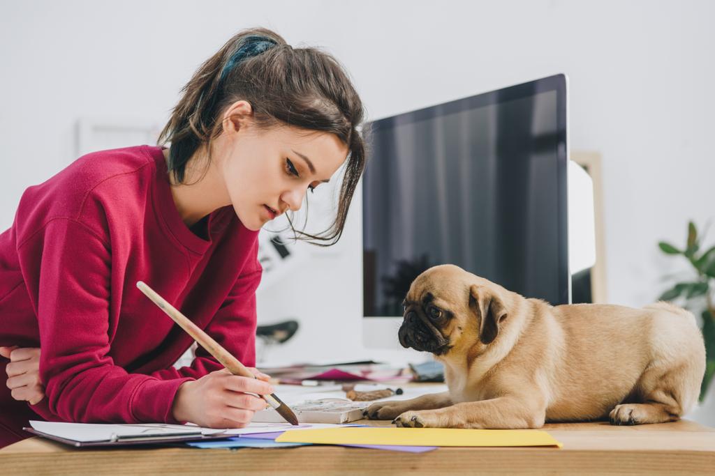 Cute pug looking at girl working on illustrations on working table with computer - Photo, Image