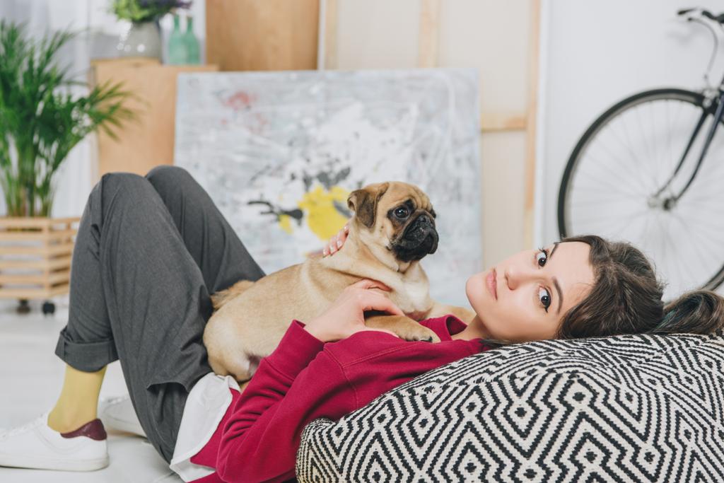 Pretty lady playing with pug puppy on large cushion - Photo, Image