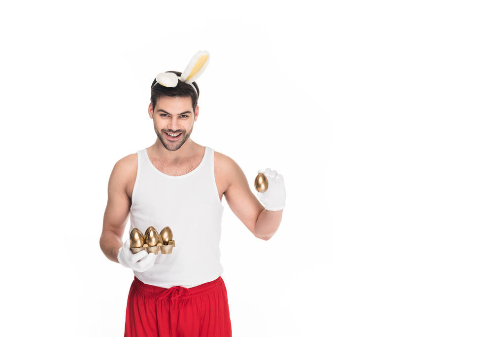 Улыбающийся человек с кроличьими ушами держит золотые яйца изолированные на белом, пасхальная концепция
 - Фото, изображение