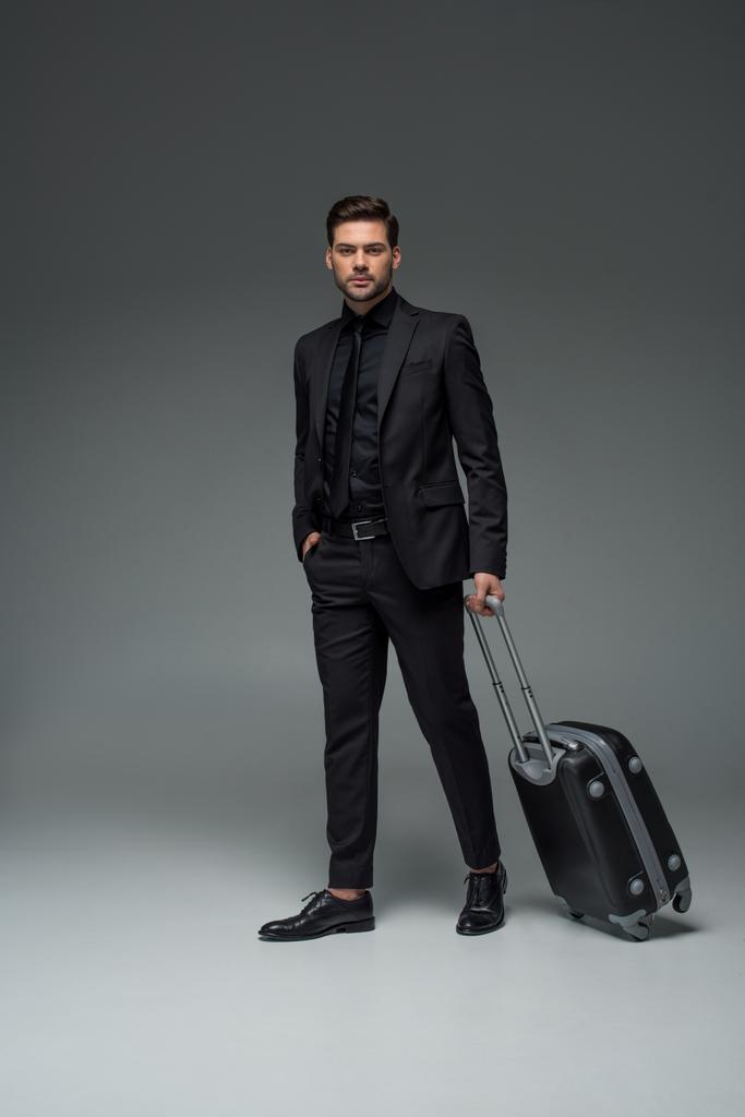 Молодий стильний турист, що носить валізу на сірому
 - Фото, зображення