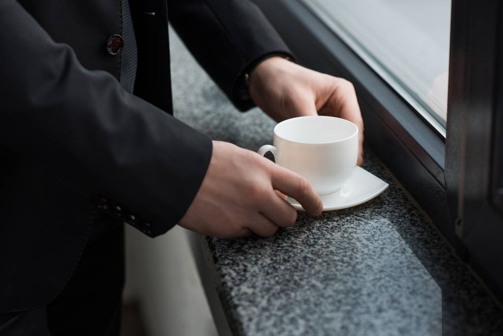 Vista parziale dell'uomo d'affari che tiene una tazza di caffè sul davanzale della finestra
 - Foto, immagini