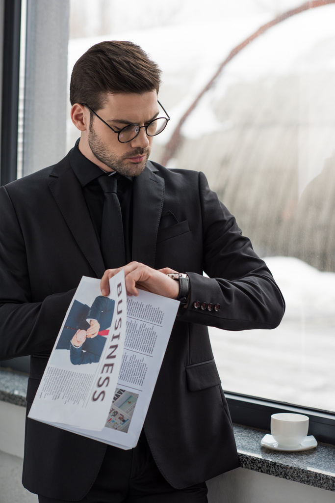 Jungunternehmer mit Brille blickt auf Armbanduhr und hält Zeitung in der Hand - Foto, Bild